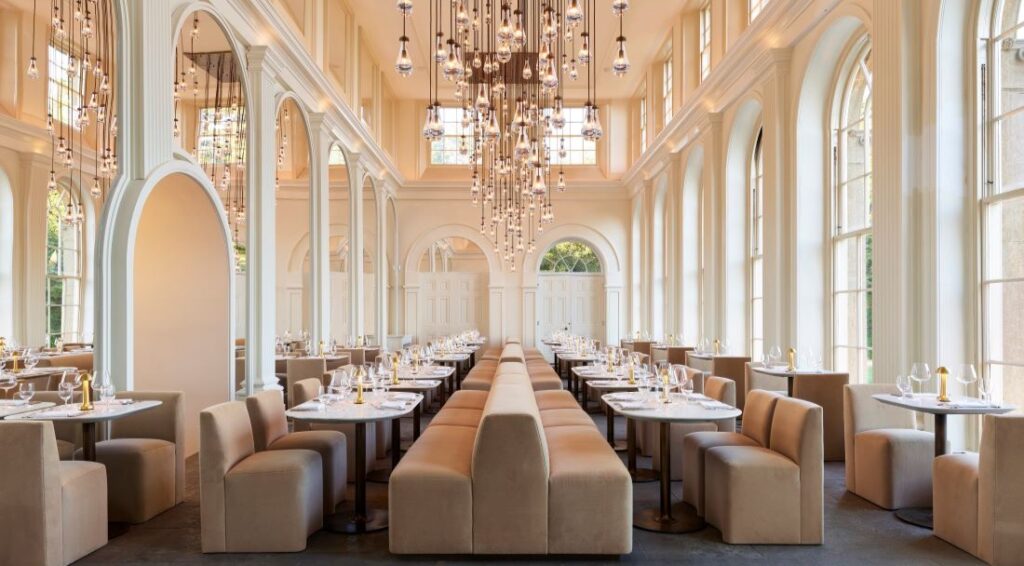 A high-ceilinged white room with rows of pale brown banquettes and chairs around white tables