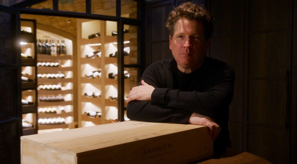 Image of winemaker Olivier Bernstein, leaning on a table, his wine cellar visible in the background